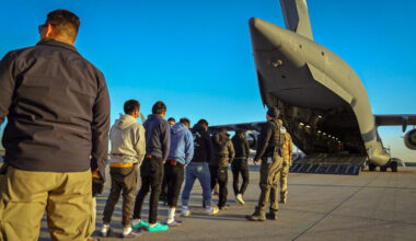 A line of men in chains, seen from behind, are marched by law enforcement officers toward the open back hatch of a massive transport aircraft.