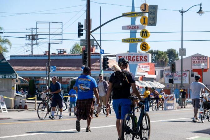 Cyclists and pedestrians walk and ride down a closed off street past an intersection with a Jims Burgers restaurant on the corner.