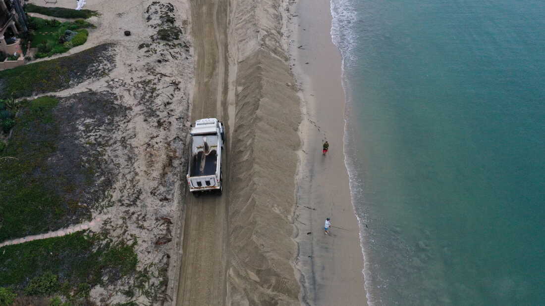 People fish in the Pacific Ocean as a dump truck unloads sand to build berms on the beach to protect homes as they prepare for elevated surf, coastal flooding, and beach erosion ahead of Hurricane Polo in Long Beach, California.