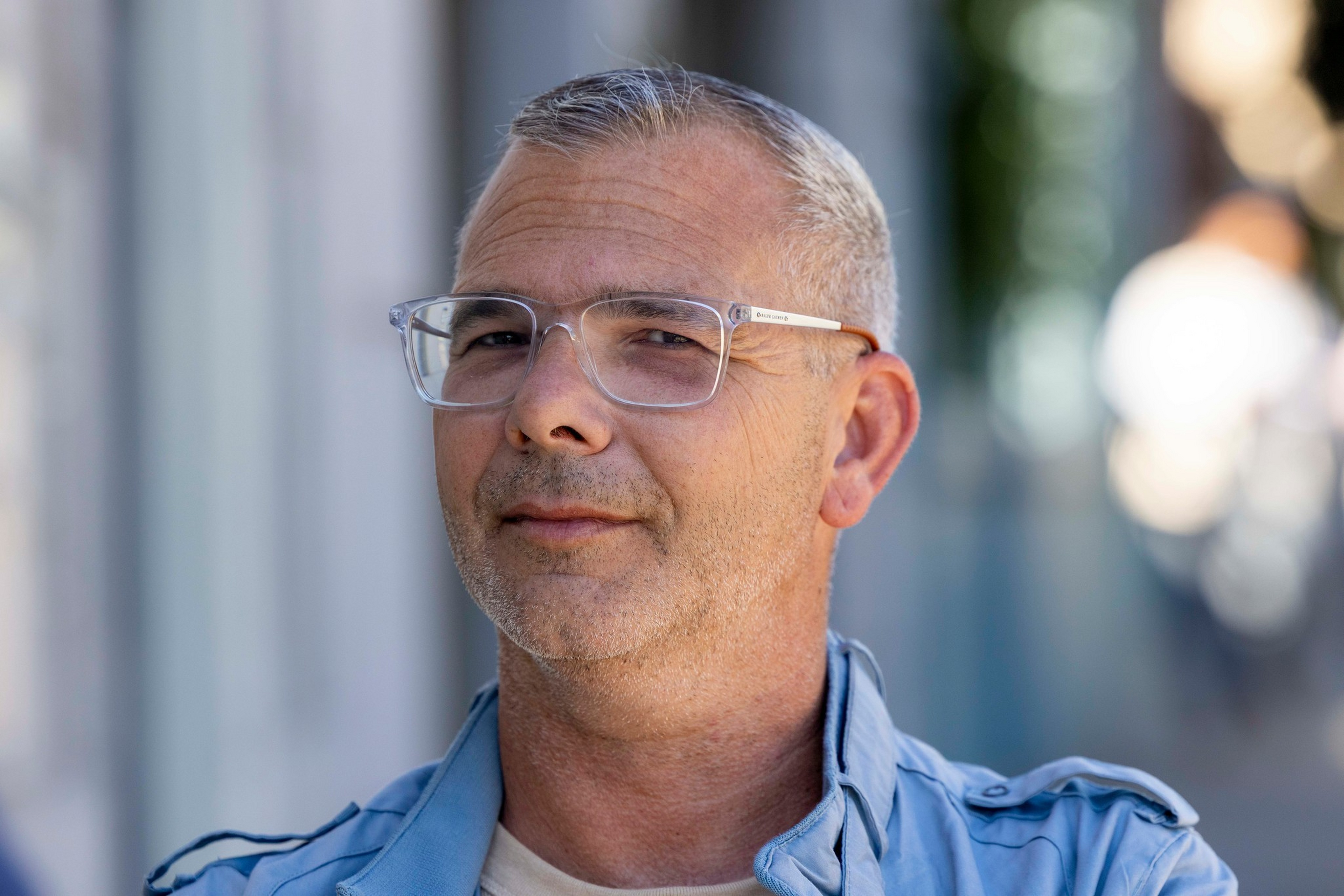 A middle-aged man with short gray hair and clear glasses wears a light blue jacket, looking slightly to the side with a calm expression.