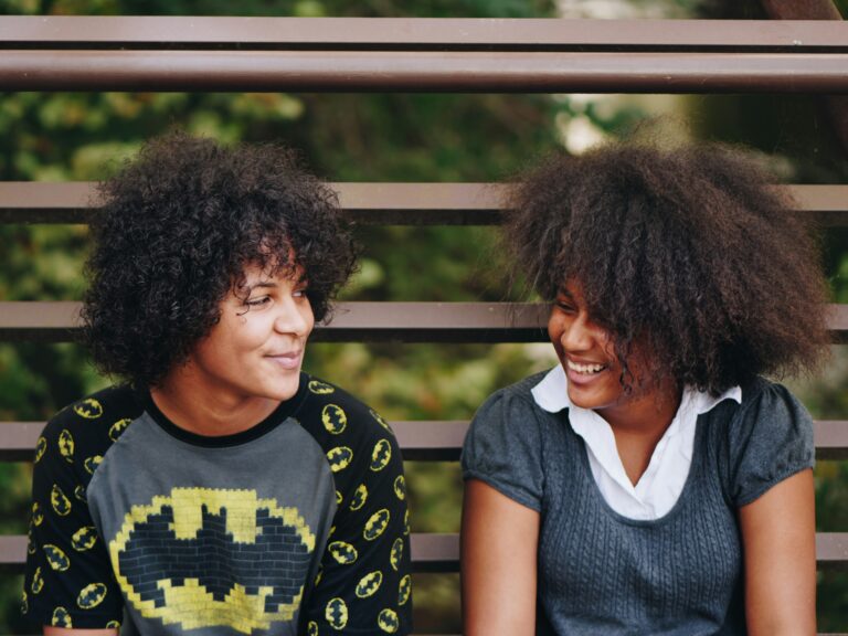 Two Black teens lean against a metal fence and smile at each other