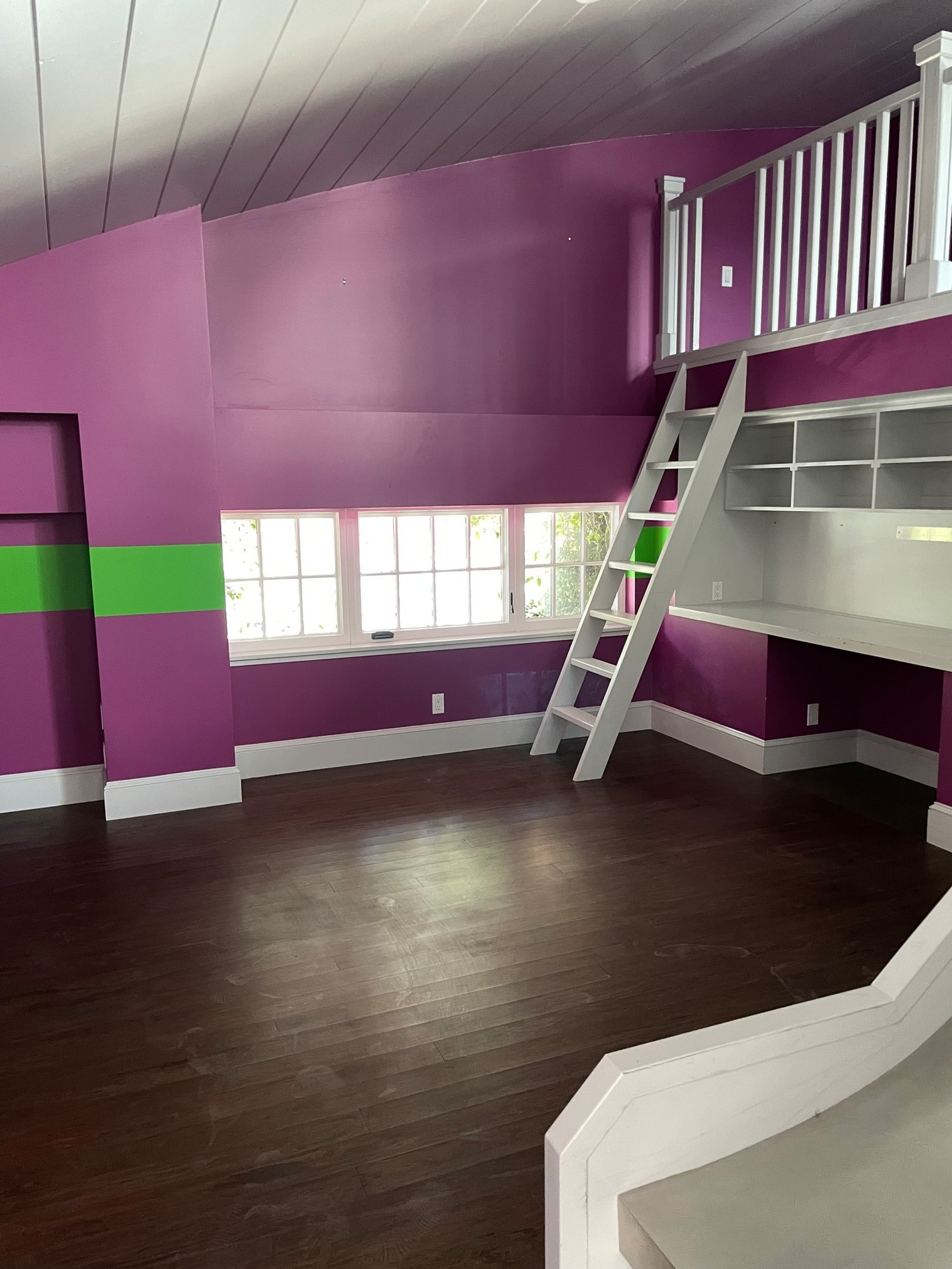 A purple room with a green stripe, dark wood floor, white loft ladder, built-in white desk with shelves, and a set of windows near the floor.