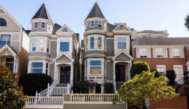 Two Victorian-style homes with pointed turrets, large windows, and front porches stand side by side, flanked by brick buildings and greenery.