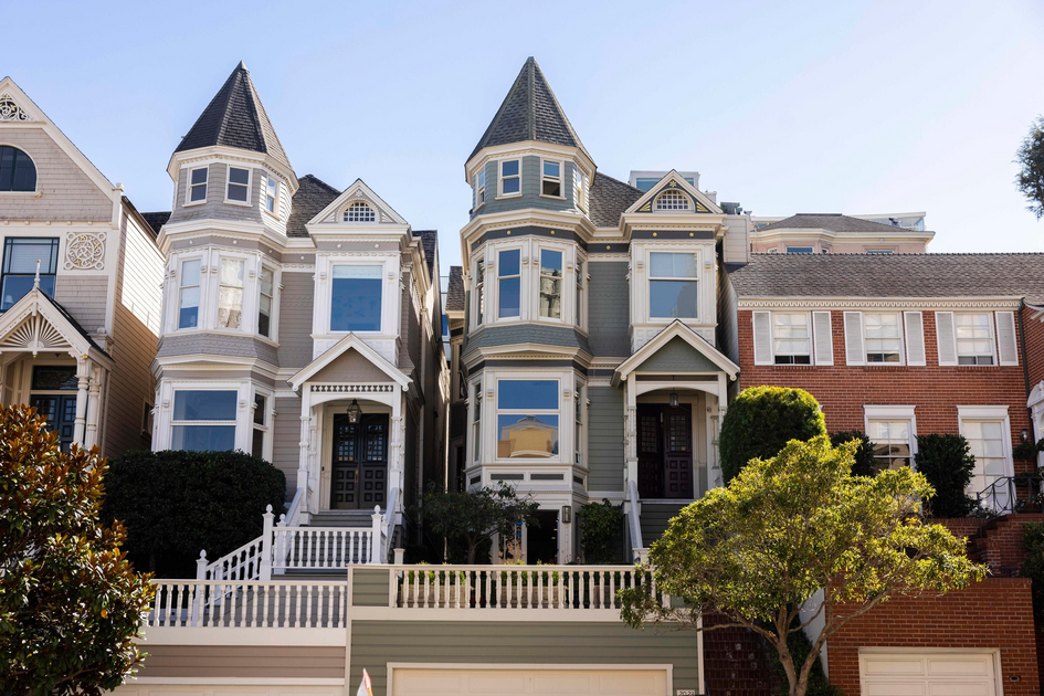 Two Victorian-style homes with pointed turrets, large windows, and front porches stand side by side, flanked by brick buildings and greenery.