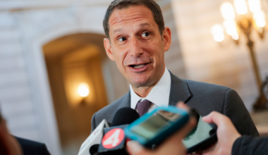 A man in a suit and tie speaks animatedly to reporters, with microphones and phones held up close to capture his statements.