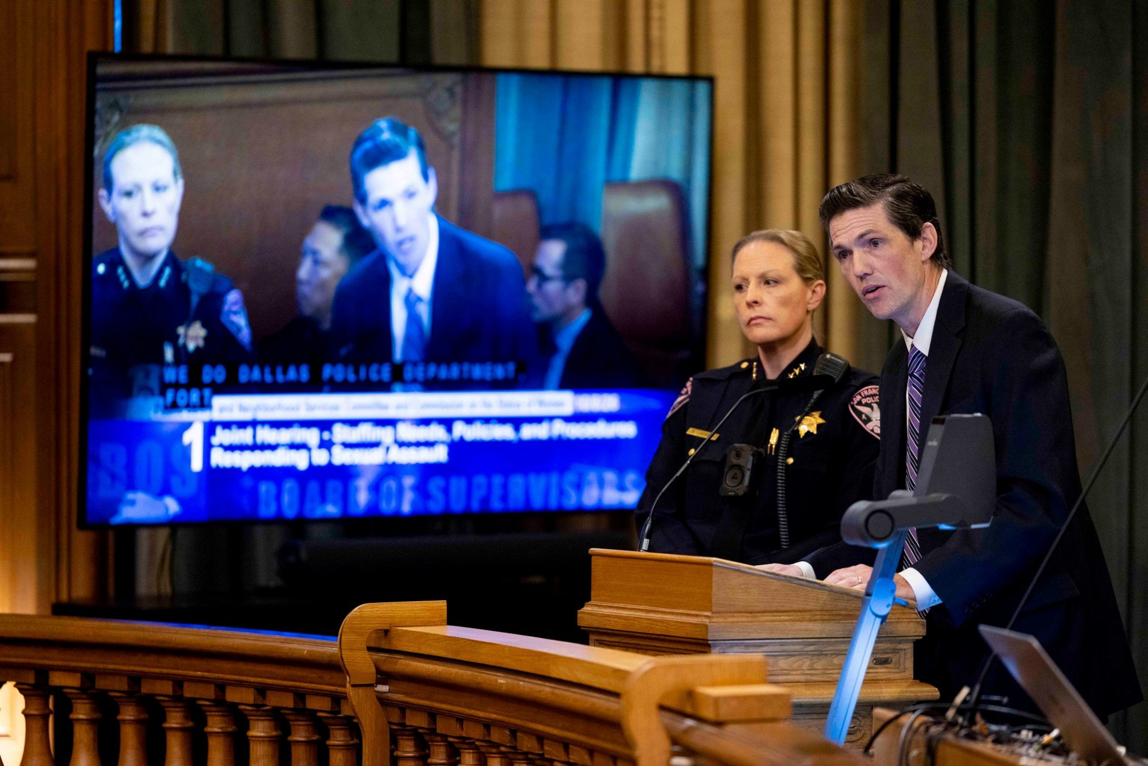 A man in a suit and a female police officer stand at a podium speaking to a board, with a large screen showing their live image behind them.
