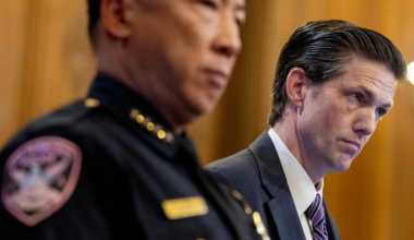 A police officer in uniform and a man in a suit with a striped tie stand side by side, both looking serious and focused.