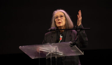 Anne Carson speaks at the 2024 National Book Awards finalist reading, standing behind a clear lectern with one hand raised.