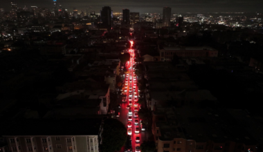 A long line of cars with red brake lights forms a nighttime traffic jam on a city street surrounded by dark buildings and a distant city skyline.
