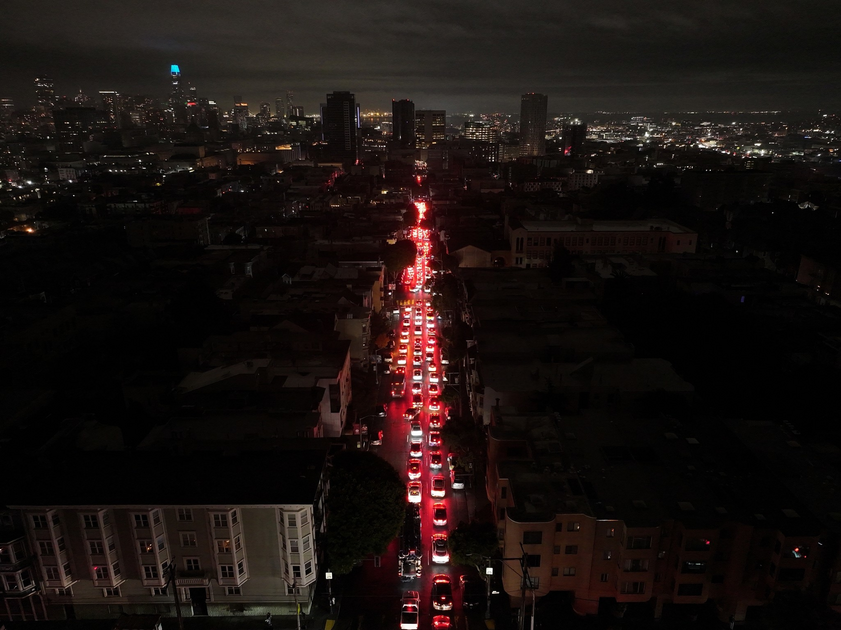 A long line of cars with red brake lights forms a nighttime traffic jam on a city street surrounded by dark buildings and a distant city skyline.