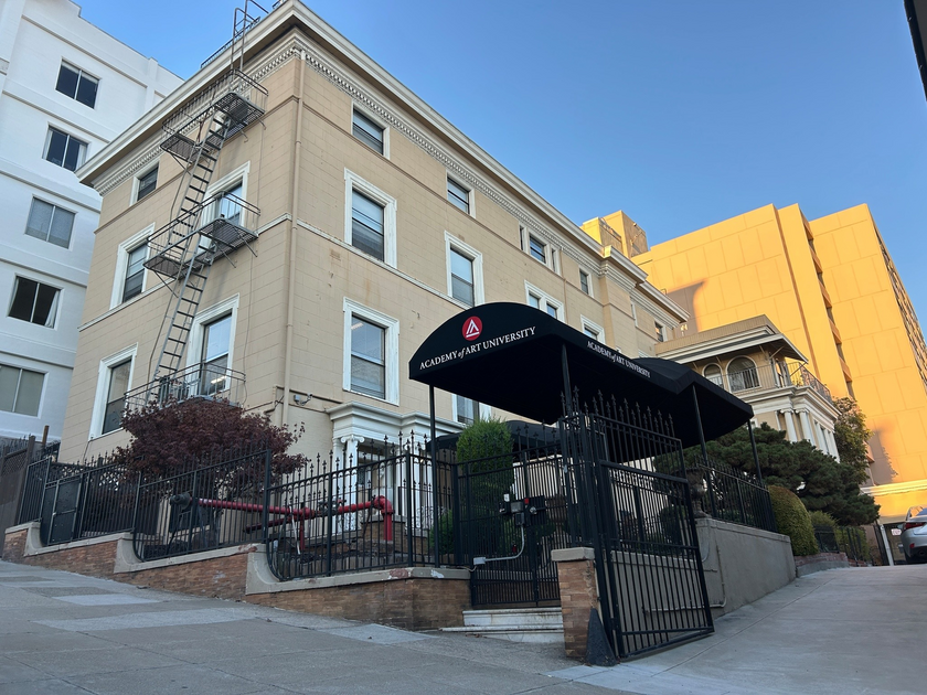 A beige three-story building with a black wrought iron fence and gate, a fire escape, and a black awning labeled "Academy of Art University."