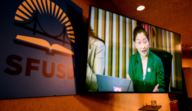 A woman in a green blazer speaks into a microphone, shown on a large screen next to a blue sign with an orange bridge and book logo labeled "SFUSD."