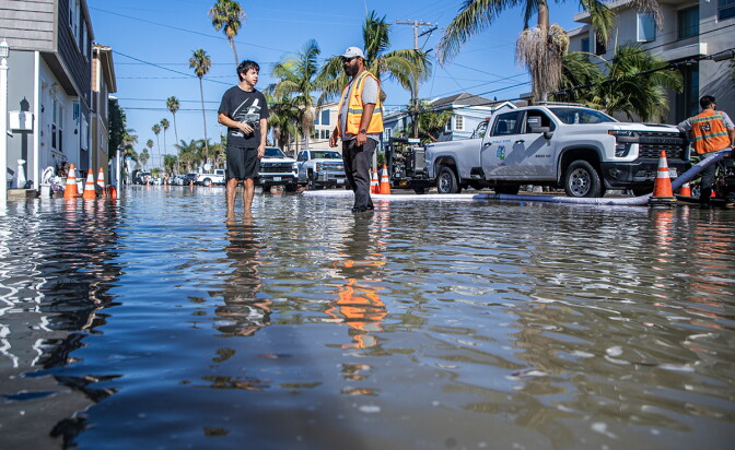 People wade through a flooded street lined with palm trees as a public works truck and crew with hoses work nearby.