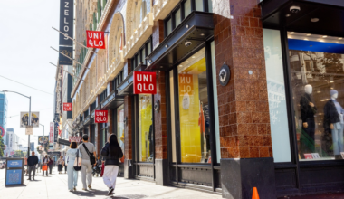 A sunny city sidewalk with several people walking past a UNIQLO store, featuring large windows and red UNIQLO signs hanging from the building.