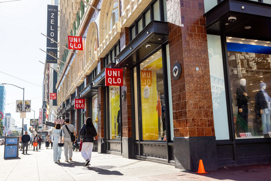 A sunny city sidewalk with several people walking past a UNIQLO store, featuring large windows and red UNIQLO signs hanging from the building.