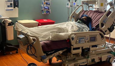 A patient lies in a hospital bed covered with a white blanket, wearing a neck brace and connected to medical monitors and IV drips.