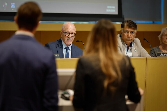Two people stand with their backs to the camera, facing a wooden dais. Behind it, a person with glasses and a light skin tone speaks into a microphone, while a person with a medium-brown skin tone, wearing a light gray blazer, looks on.