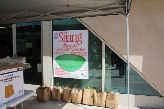 Large signage is post on a window that reads "Sijang. Mutual aid and community marketplace" next to bags of produce.