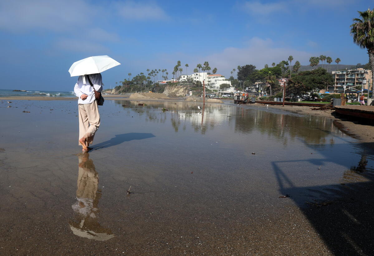 A woman in a light colored outfit carrying a light colored umbrella is reflected in water at the beach.