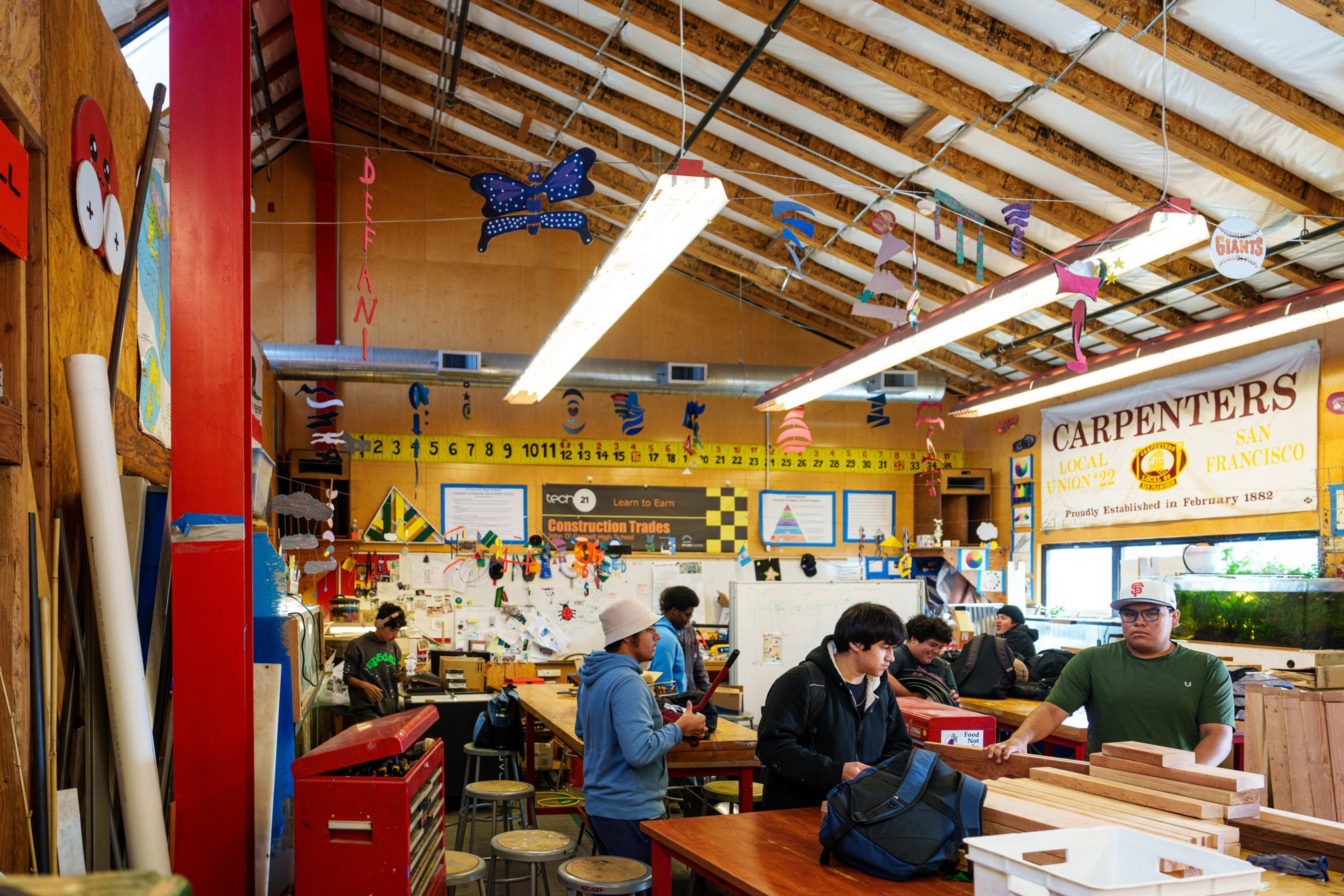 A woodworking classroom with students working, colorful crafts hanging from the ceiling, and a large “Carpenters Local Union 22" banner on the wall.