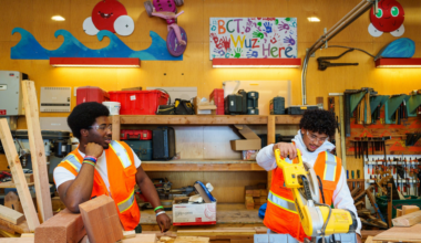 Two young men in orange safety vests work in a woodshop surrounded by wooden planks, tools, and colorful wall decorations with waves and a sun.