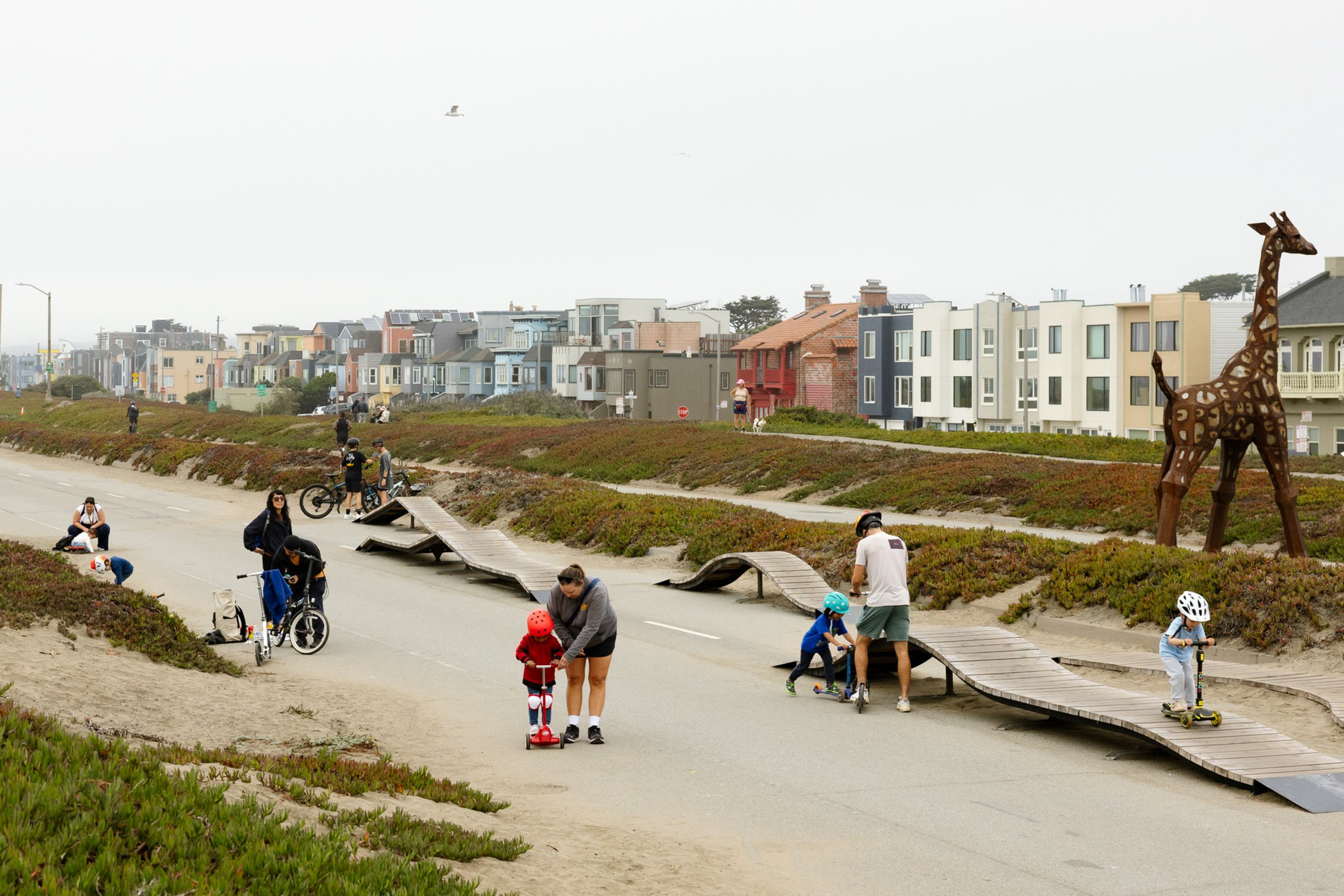 Children and adults ride scooters and bikes on a path with wooden ramps near residential houses and a large giraffe sculpture.