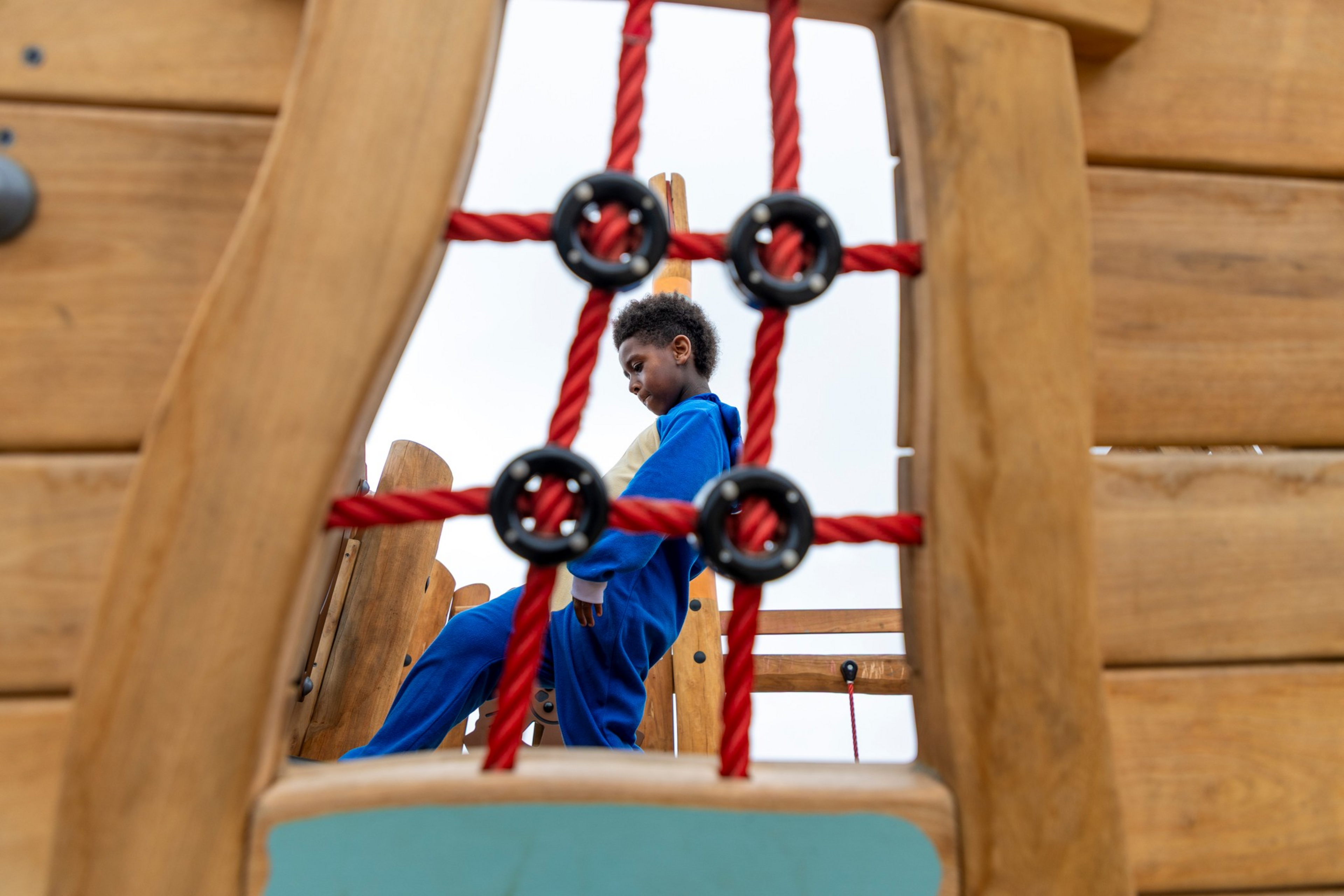 A child in blue clothes climbs on a wooden playground structure, seen through a red rope net with black circular connectors.