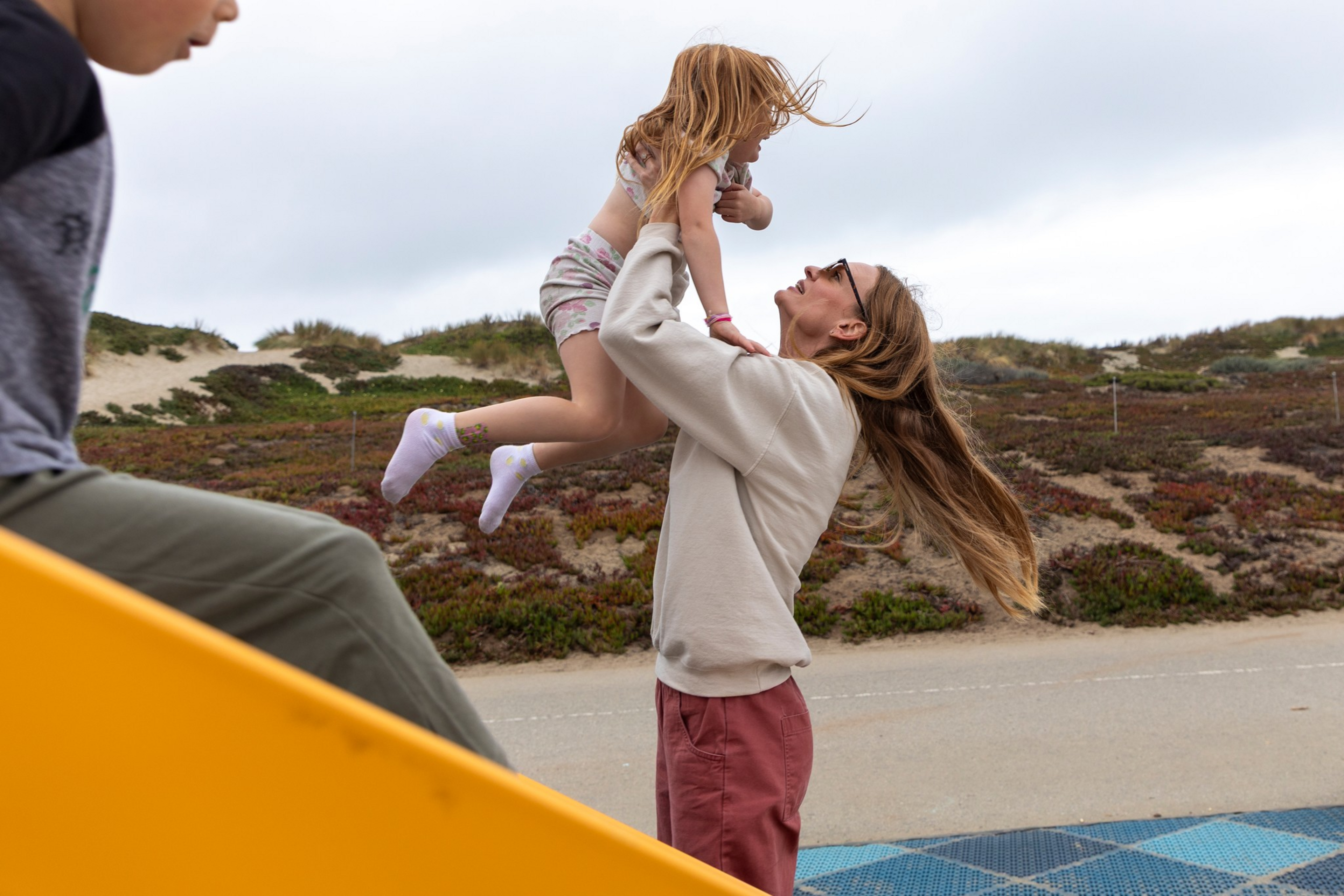 A woman lifts a young girl into the air at a playground, with a boy sitting nearby on a yellow slide and a grassy hill in the background.