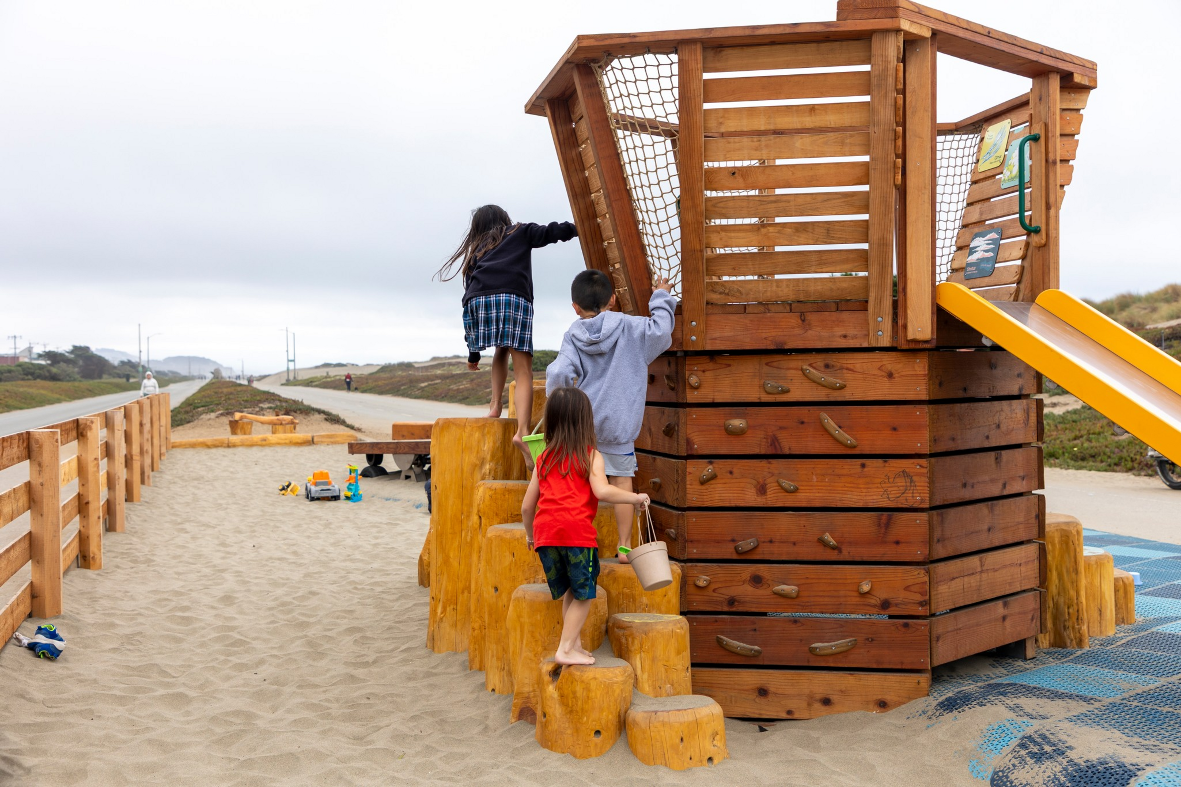 Three children climb a wooden play structure resembling a ship, with sand beneath and a yellow slide on the right side.