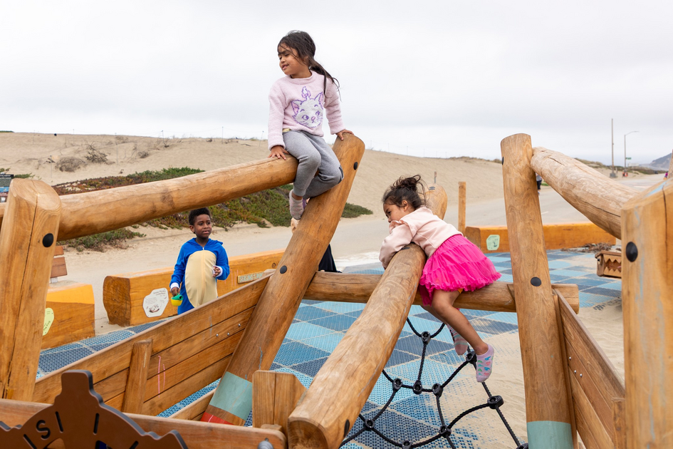 Two girls climb on a wooden playground structure, one sitting on top, another leaning over a beam, while a boy watches nearby on a cloudy day.