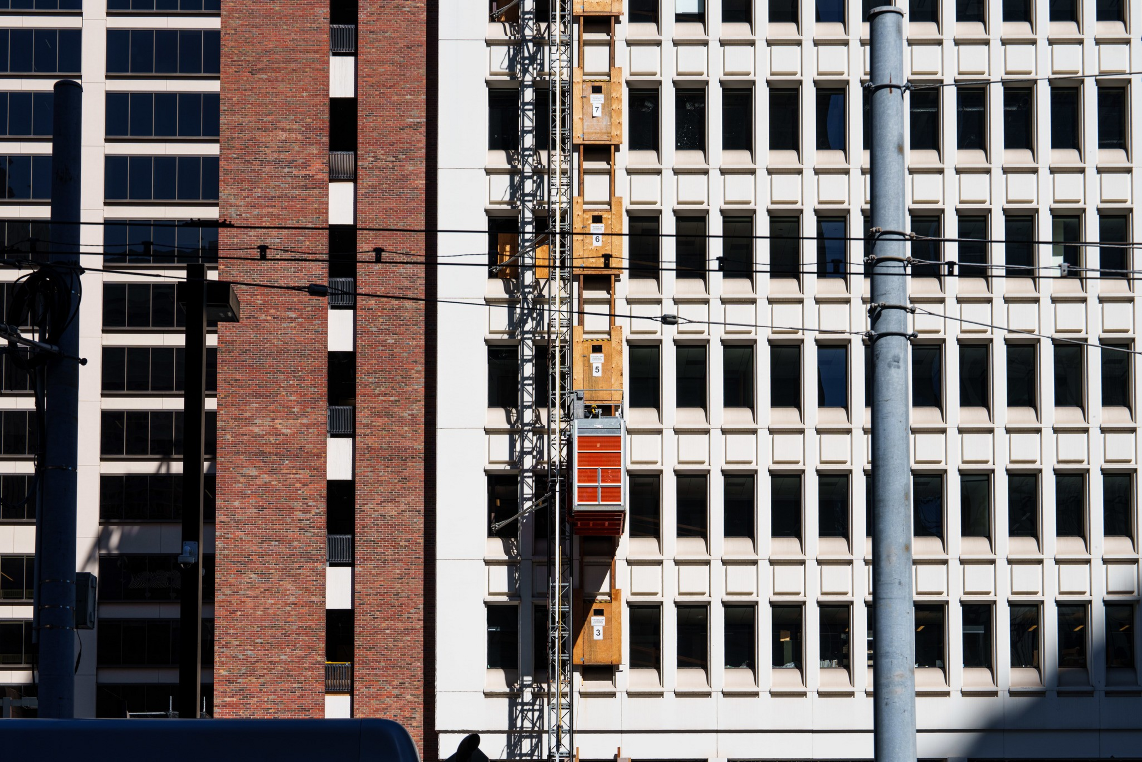A red construction elevator is attached to the side of a white office building with grid-like windows, next to a brown brick building.