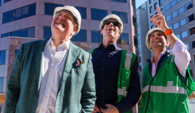 Three men wearing safety helmets and green vests stand outdoors in front of tall buildings, smiling and looking up, with one man pointing upwards.