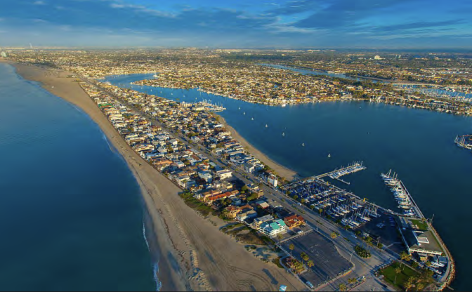An aerial view looking down a long, narrow strip of land packed with homes and buildings, with a wide sandy beach and open ocean on the left and a calm bay on the right.