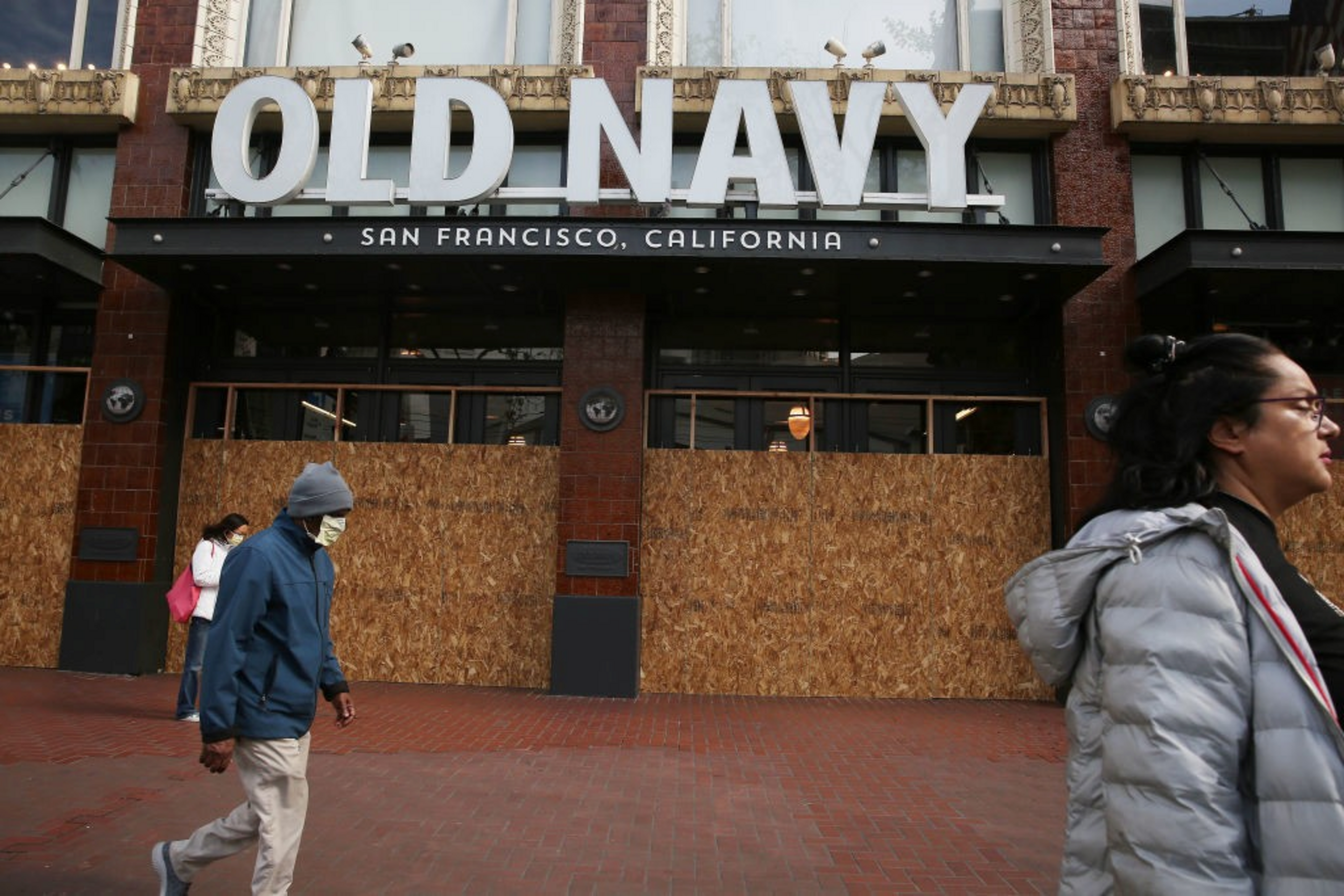 Old Navy store in San Francisco has its windows boarded up with plywood while three people wearing jackets and masks walk past.
