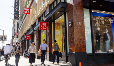 People walk past a Uniqlo store on a sunny city sidewalk, with mannequins and bright displays visible through large windows.