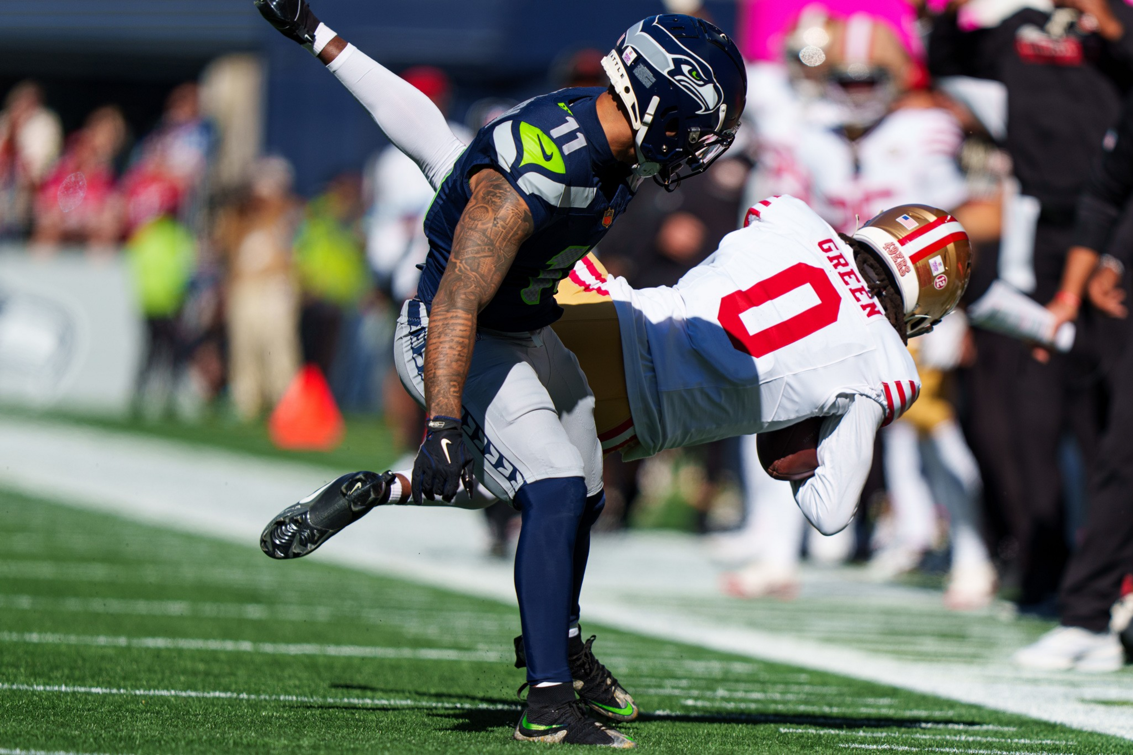 A Seattle Seahawks player tackles a San Francisco 49ers player wearing number 0 who is bent over while holding the football near the sideline.