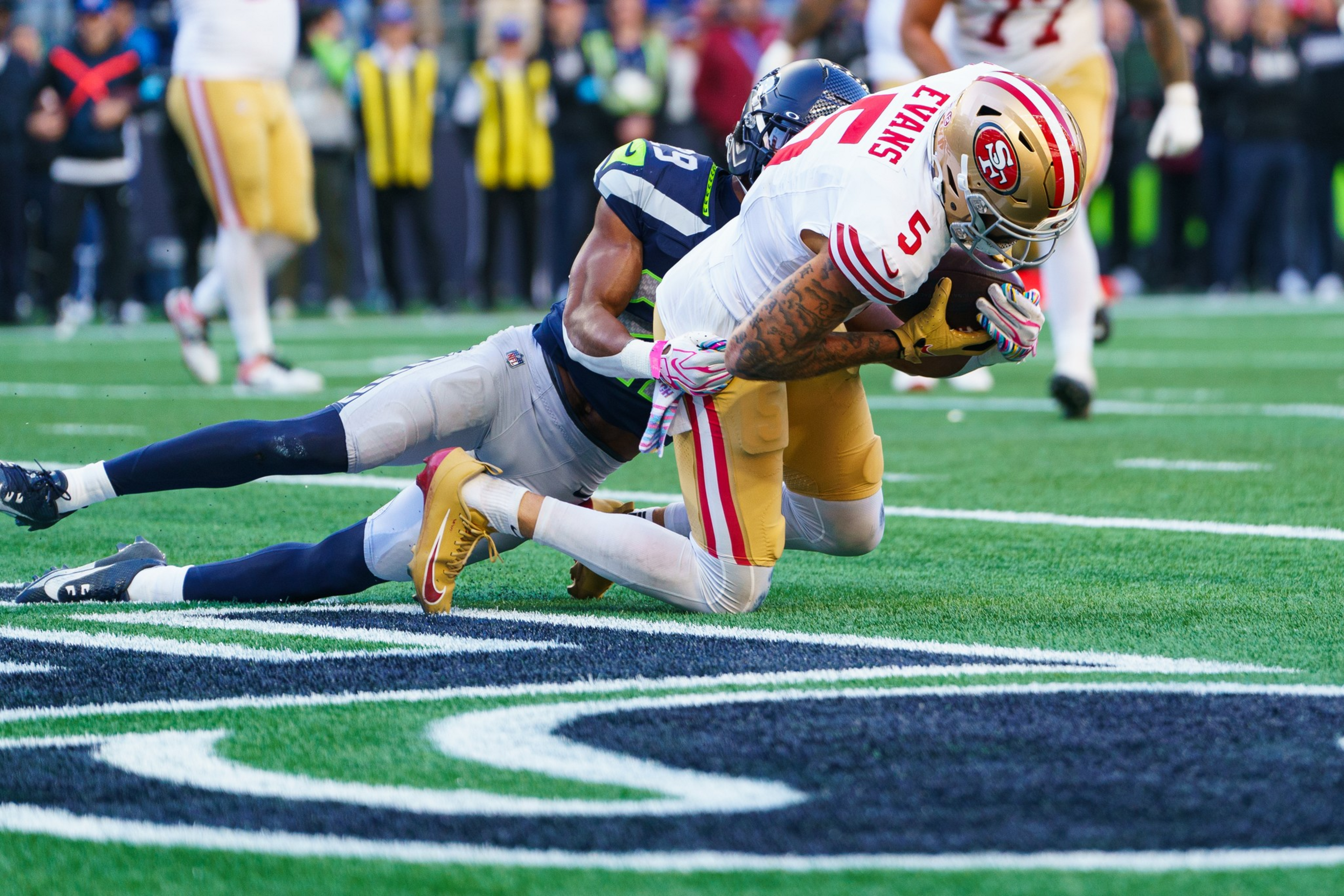 A San Francisco 49ers player wearing number 5 dives forward while being tackled by a Seattle Seahawks defender near the end zone.