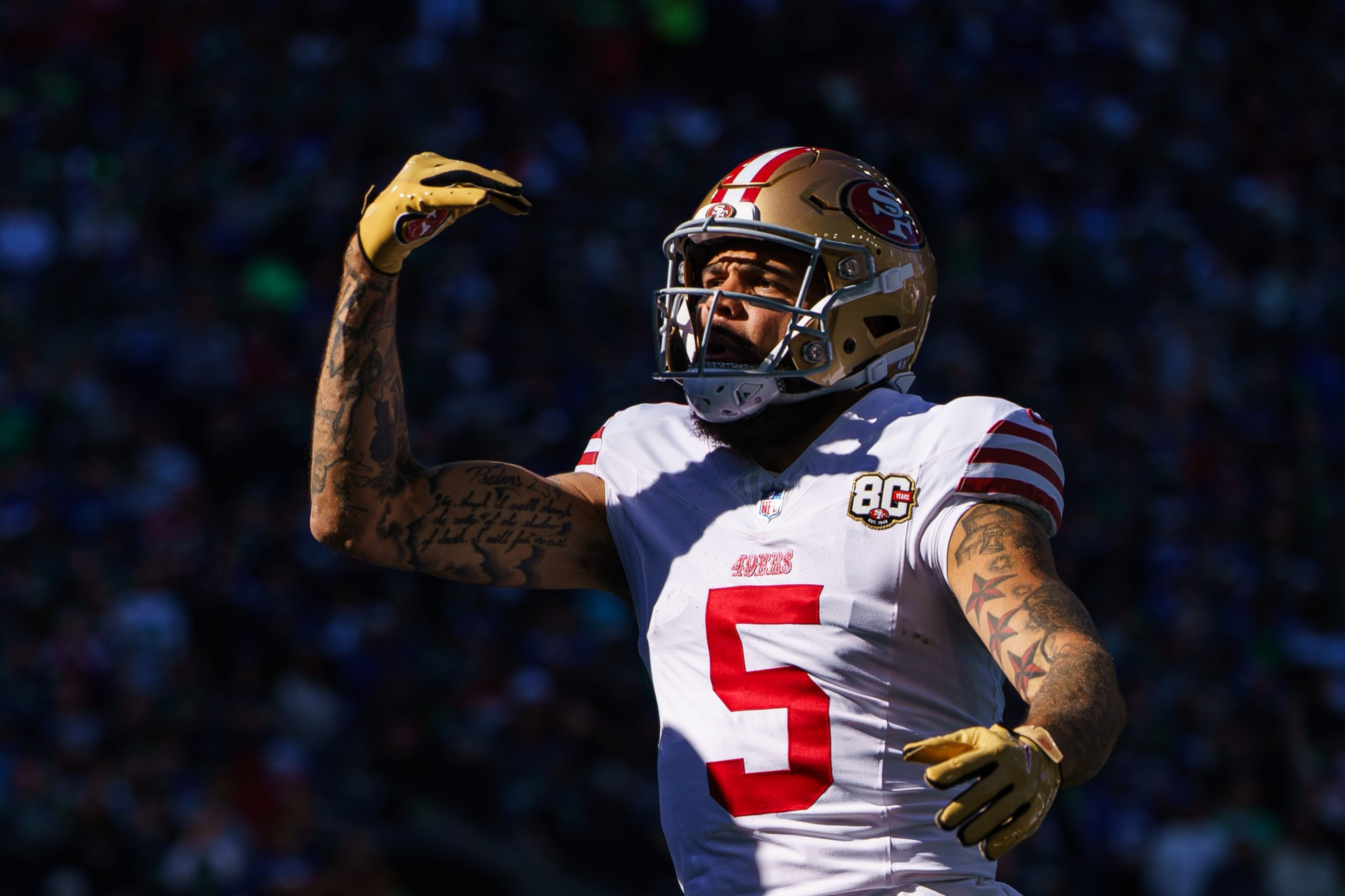 A football player in a white 49ers jersey with the number 5 raises his right arm, wearing a gold helmet and gloves, with visible tattoos on both arms.