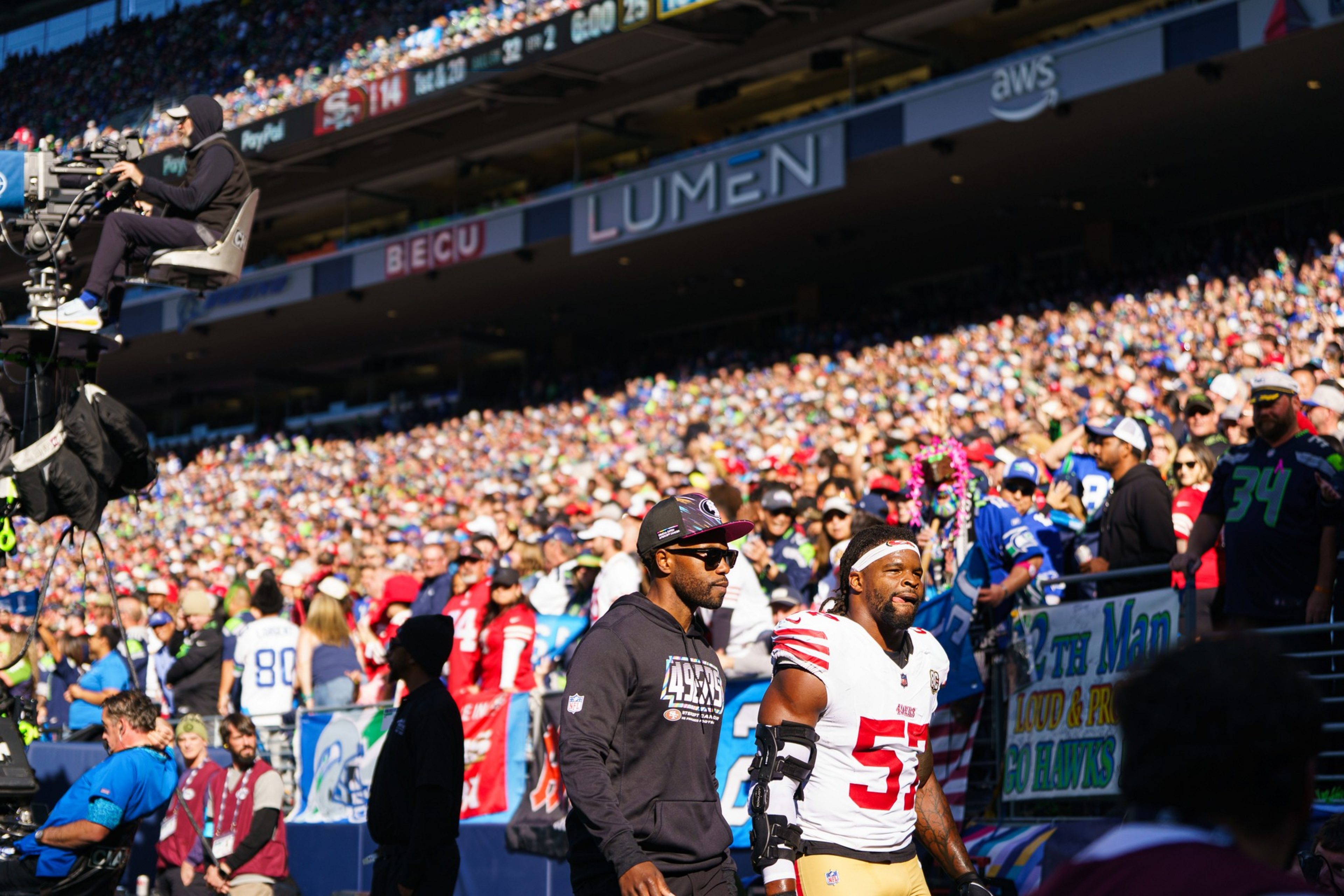 Two NFL players walk along the sideline during a game with a crowded stadium and a cameraman filming from above.