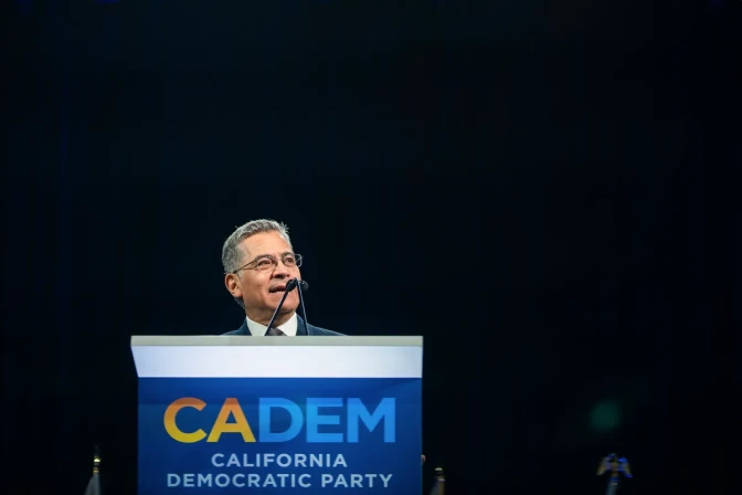 A low angle view of Xavier Becerra, a man with medium skin tone, speaking in front of a podium with signage on it that reads "CADEM. California Democratic Party."