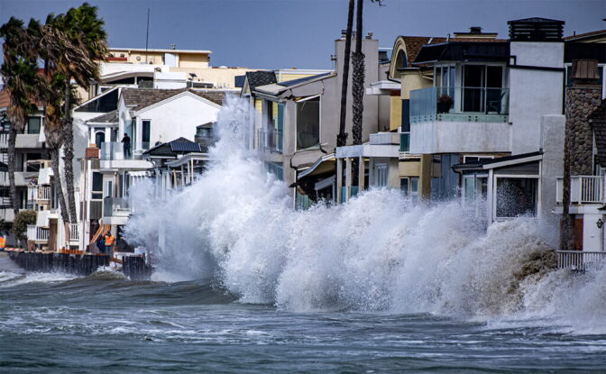 A massive wave breaks against a tightly packed row of multistory beachfront homes, throwing white foam and spray as high as the second floors. Dark, choppy water fills the foreground.