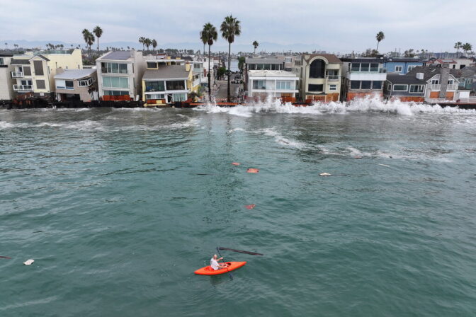 An aerial view of a kayaker in the ocean watching waves crashes into a small boardwalk and homes right in front of it.