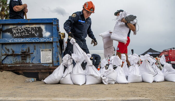 People fill sandbags on a beach. One person wears dark blue overalls and an orange helmet.