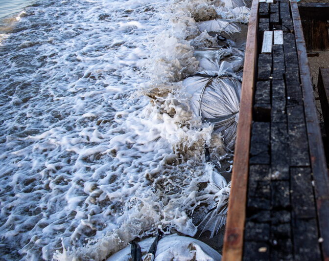 Waves crash into sand bags next to a seawall.