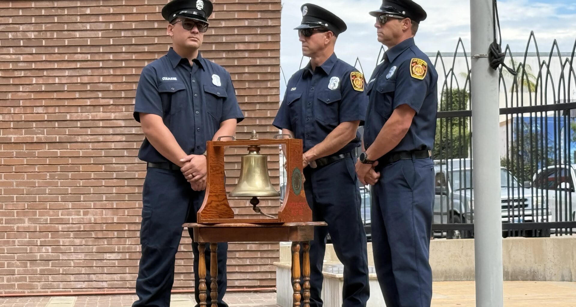 Three LAFD firefighter stand with their arms folded in from of them surrounding a brass bell mounted in a wooden case