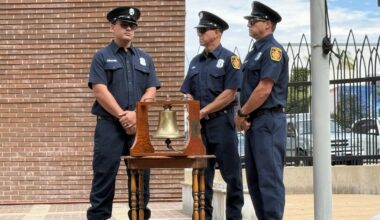 Three LAFD firefighter stand with their arms folded in from of them surrounding a brass bell mounted in a wooden case