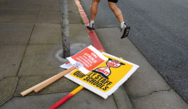 Two protest signs lie on the sidewalk near a red curb while a person wearing shorts and sandals steps off the curb onto the street.