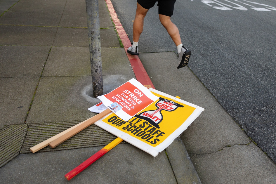 Two protest signs lie on the sidewalk near a red curb while a person wearing shorts and sandals steps off the curb onto the street.