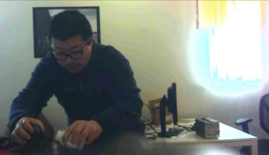 A person wearing glasses and a dark jacket pours liquid from a bottle into a container while sitting at a table in a dimly lit room.