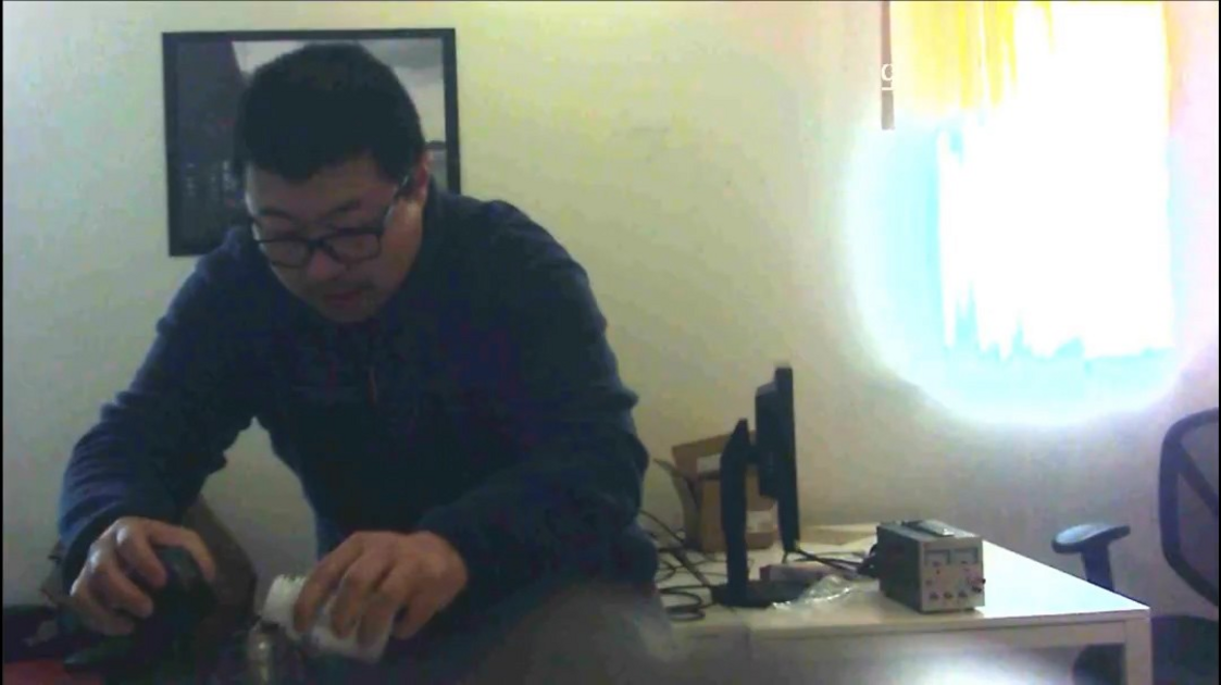 A person wearing glasses and a dark jacket pours liquid from a bottle into a container while sitting at a table in a dimly lit room.